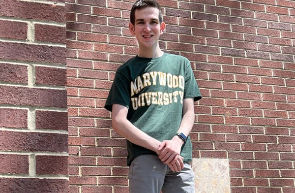 A young man wearing a Marywood University T-shirt in front of a brock wall.